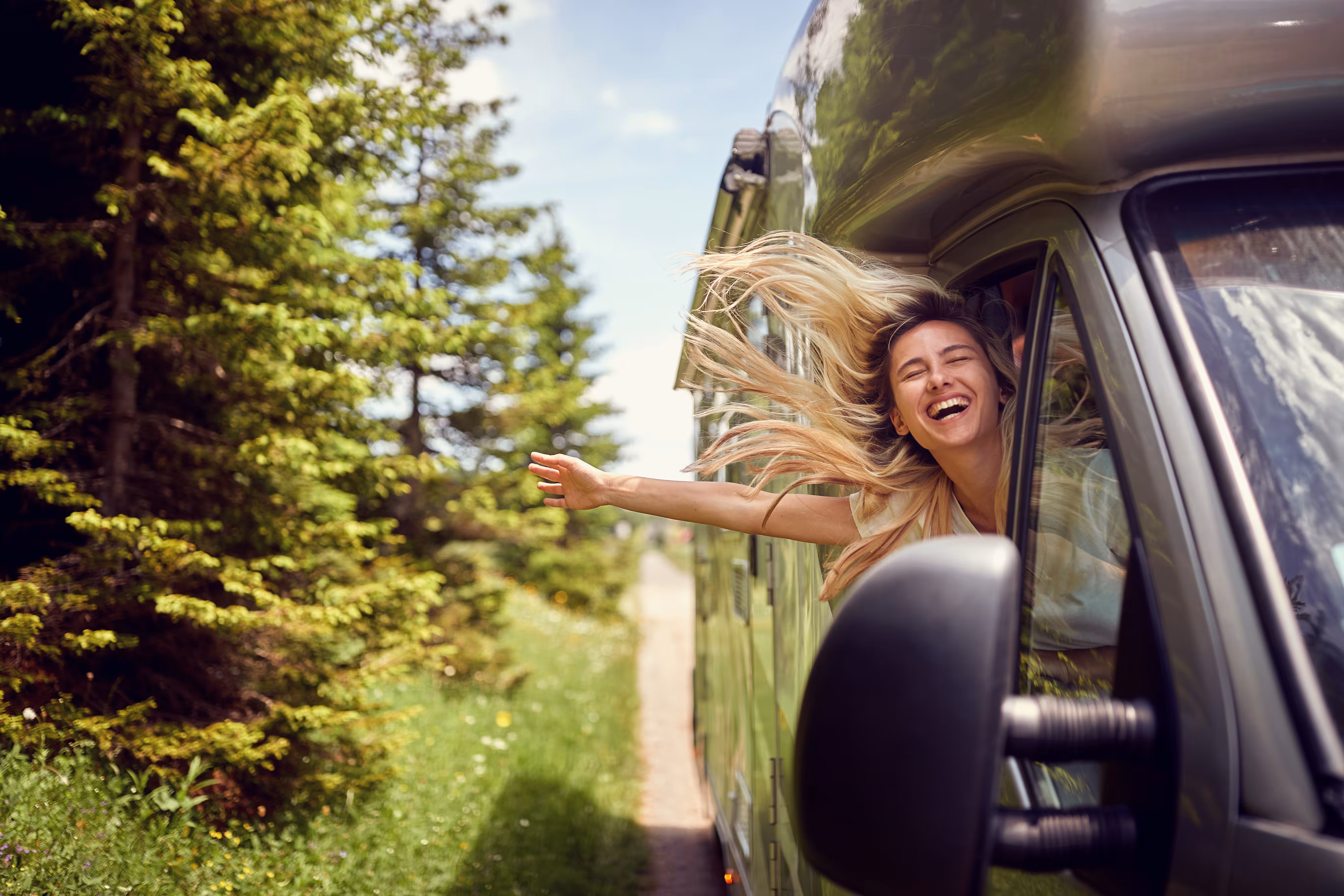 A woman with her hand out a motorhome driving down a forest road
